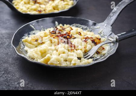 Der traditionelle schwäbische Käsespätzle mit gebratenen Zwiebeln wurde als Nahaufnahme in einer gusseisernen Bratpfanne auf einem rustikalen schwarzen Brett serviert Stockfoto