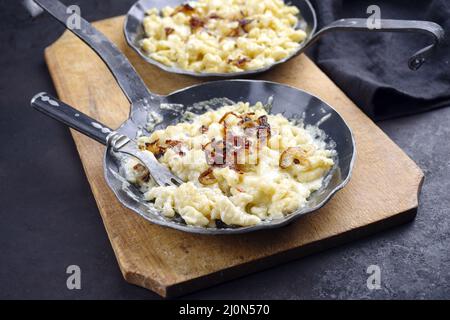 Der traditionelle schwäbische Käsespätzle mit gebratenen Zwiebeln wurde als Nahaufnahme in einer gusseisernen Bratpfanne auf einem rustikalen schwarzen Brett serviert Stockfoto