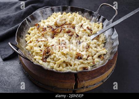 Der traditionelle schwäbische Käsespätzle mit gebratenen Zwiebeln wurde als Nahaufnahme in einer gusseisernen Bratpfanne auf einem rustikalen schwarzen Brett serviert Stockfoto