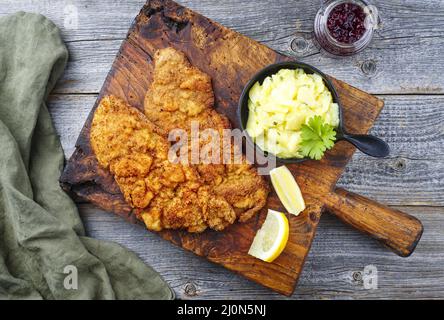 Traditionelles frittiertes Schnitzel mit Kartoffelsalat und Preiselbeeren serviert als Draufsicht auf einem alten rustikalen Holzbrett Stockfoto