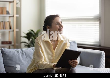 Eine fröhliche, nachdenkliche Dame mittleren Alters, die lächelnd auf dem Sofa sitzt Stockfoto