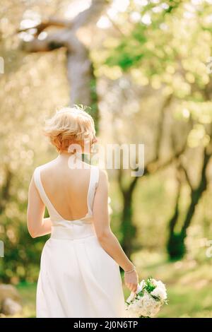 Die Braut in einem weißen Kleid mit einem Blumenstrauß in den Händen steht im Park mit dem Kopf nach rechts gedreht Stockfoto