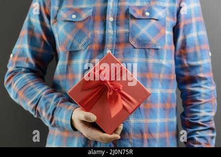 Eine rote Geschenkbox mit einer Schleife in der Hand eines Mannes in einem blauen karierten Hemd. Festliche Überraschung Stockfoto