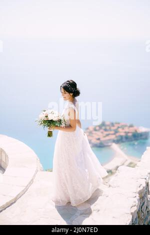 Die Braut mit Brautstrauß steht auf der Aussichtsplattform mit Blick auf die Insel Sveti Stefan, Rückansicht Stockfoto