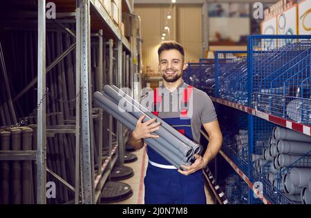 Fröhlicher junger Verkäufer, der in einem der Gänge des Ladens steht und PVC-Abflussrohre hält Stockfoto