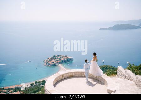 Die Braut steht auf der Brüstung der Aussichtsplattform mit Blick auf die Insel Sveti Stefan, der Bräutigam hält ihre Hand Stockfoto