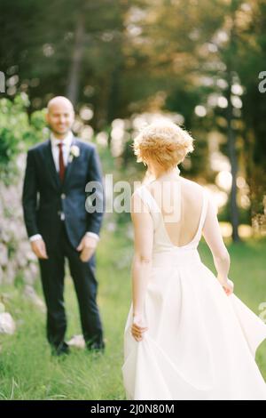 Die Braut in einem weißen Kleid geht vor dem Hintergrund grüner Bäume zum lächelnden Bräutigam zu Stockfoto