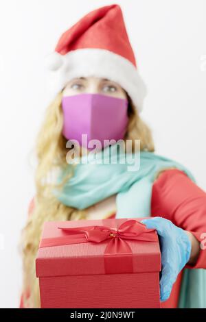 Eine Schachtel mit einem Weihnachtsgeschenk in der Hand des Mädchens. Eine Frau in einem Weihnachtsmann Hut in einer medizinischen Maske und Handschuhen. Voller Schutz für Stockfoto