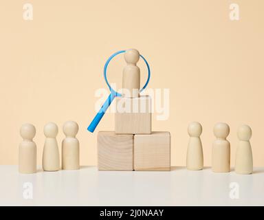 Holzfiguren von Männern stehen auf einem beigen Hintergrund und einer blauen Plastiklupe. Rekrutierungskonzept, Suche nach talentierten Stockfoto