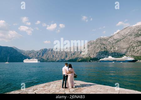 Braut und Bräutigam umarmen sich am Pier vor dem Hintergrund von Linern und Bergen. Rückansicht Stockfoto