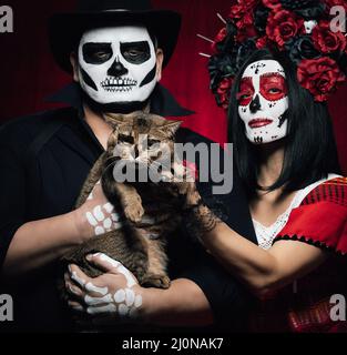 Schöne Frau mit einem Zuckerschädel Make-up mit einem Blumenkranz auf ihrem Kopf und einem Skelett Mann in einem schwarzen Hut mit einem grauen Stockfoto