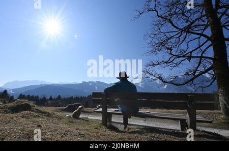 Garmisch Partenkirchen, Deutschland. 20. März 2022. Ein Spaziergänger mit Hund ruht sich auf einer Bank in der Sonne aus. Quelle: Angelika Warmuth/dpa/Alamy Live News Stockfoto