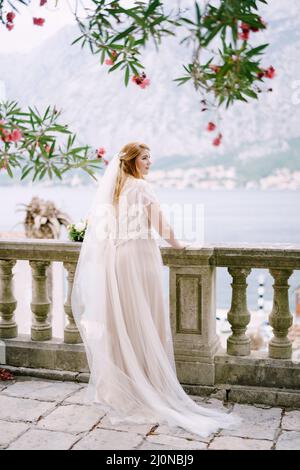 Braut steht auf einer Steinterrasse in der Nähe der Balustrade und blickt auf das Meer Stockfoto