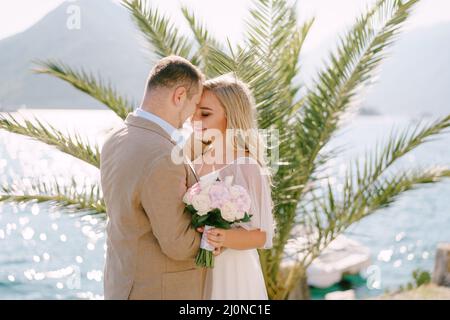 Der Bräutigam und die lächelnde Braut mit einem Blumenstrauß stehen auf dem Pier im Hintergrund einer Palme und umarmen sich zärtlich Stockfoto