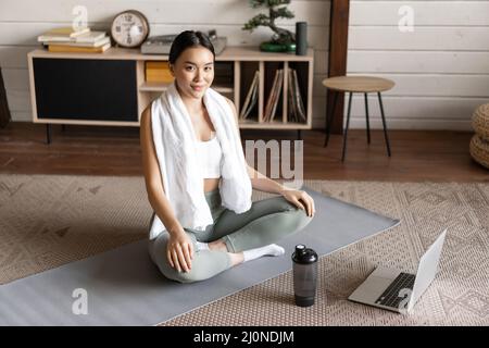 Asiatische Frau, die zu Hause Morgenyogas macht, in Meditationspose auf der Matte sitzt, aktive Beinkleider trägt und friedlich lächelt Stockfoto