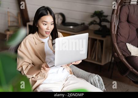 Asiatische Frau sieht aufgeregt und erstaunt über Laptop-Bildschirm, gewinnen online, zu Hause sitzen Stockfoto