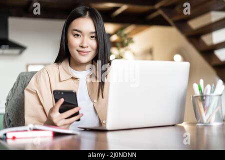 Lächelndes asiatisches Mädchen nutzt Computer zu Hause, hält Smartphone und sieht glücklich an der Kamera. Vielbeschäftigte Frau, die an einem Laptop mit Fernbedienung arbeitet Stockfoto