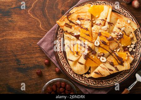 Crepes-Suzette mit Orangen und Schokolade. Dünne Crepes mit Schokoladenaufstrich, Haselnüssen und Orangenscheiben Obst auf dem Teller zum Frühstück auf altem dunklen Woo Stockfoto