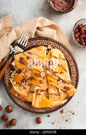 Crepes-Suzette mit Orangen und Schokolade. Dünne Crepes mit Schokoladenaufstrich, Haselnüssen und Orangenscheiben Obst auf dem Teller zum Frühstück auf altem Beton Stockfoto