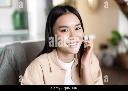 Lächelnde junge asiatische Frau, die ihr Gesicht berührt und in der gemütlichen Küche sitzt. Mädchen sitzt zu Hause und sieht glücklich an der Kamera, moderne Millenie Stockfoto