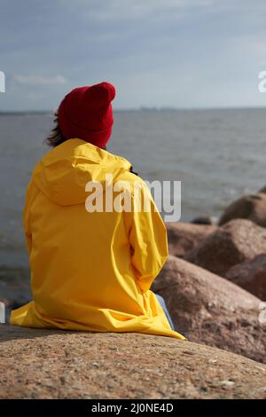 Rückansicht einer Frau, die in einem leuchtend gelben Mantel und einem roten Hut auf Stein in der Nähe der Nordseeküste sitzt Stockfoto