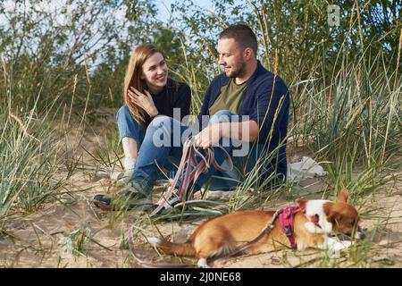 Junges glückliches Paar mit Corgi-Hund, der am Sand sitzt. Schöner Mann und schöne Frau Stockfoto