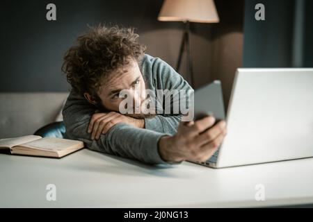 Gelangweilter Student in einem Online-Video-Kurs swipes auf seinem Telefon in sozialen Netzwerken. Der junge Mann verschwendet Zeit damit, Bilder auf zu sehen Stockfoto