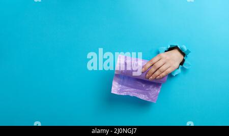 Weibliche Hand hält ein Paket transparenter Müllbeutel auf blauem Hintergrund. Ein Teil des Körpers ragt aus einem zerrissenen Loch im heraus Stockfoto