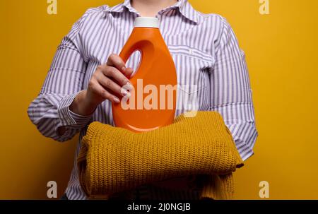 Eine Frau in einem weiß gestreiften Hemd hält eine orangefarbene Plastikflasche mit flüssigem Waschgel und einem Strickpullover. Wäscherei und Haus Stockfoto
