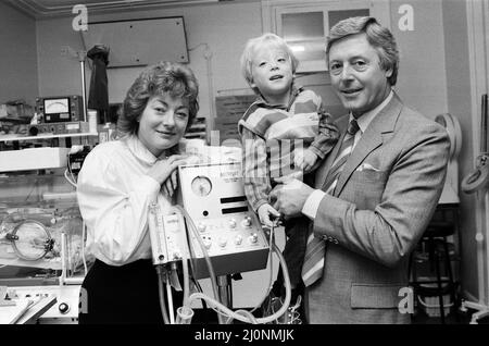 Der Fernsehpersönlichkeit Michael Aspel, der Vizepräsident von Bliss ist, kehrte ins West London Hospital zurück, um einige Geräte an die Special Care Baby Unit zu übergeben. Mit ihm waren seine Frau Lizzie Power und der 3-jährige Sohn Patrick, der vorzeitig geboren wurde und viele Wochen in der Babyeinheit verbrachte. Die von der Wohltätigkeitsorganisation gespendete Ausrüstung ist ein Beatmungsgerät – ein unverzichtbarer Bestandteil, um Neugeborenen zu helfen, die alleine nicht atmen können. Es wurde dem beratenden Kinderarzt Dr. H Barry übergeben. 3.. Oktober 1984. Stockfoto