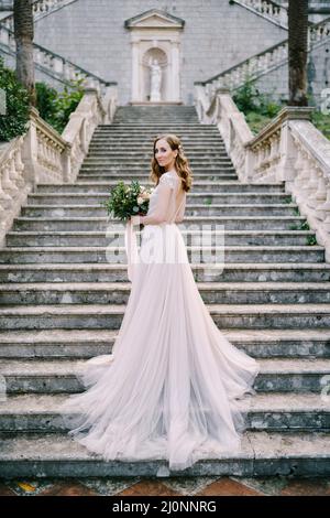 Braut in einem langen Kleid steht halb eingewickelt auf Steintreppen Stockfoto