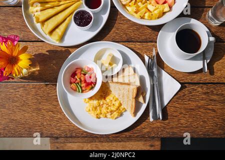 Frühstück im Touristenresort. Blick von der Tischplatte mit Rührei und Toast, Brot, Pfannkuchen, Obstsalat und Kaffee. Stockfoto