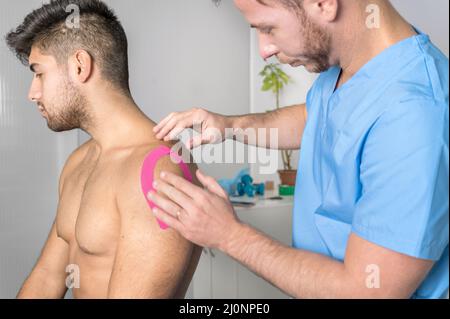 Physiotherapeut, der Kinesioband auf die Schulter des männlichen Patienten aufklebt. Kinesiologie, Physiotherapie, Rehabilitationskonzept. Schließen Stockfoto