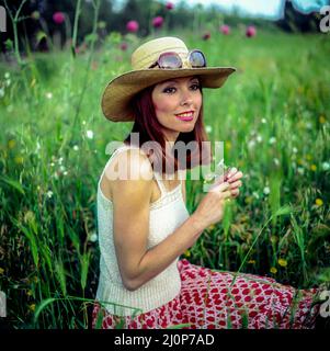 Vintage Italy 1970s, lächelndes, hübsches Frauenporträt mit Sonnenbrille auf Strohhut auf Wiese, Europa, Stockfoto