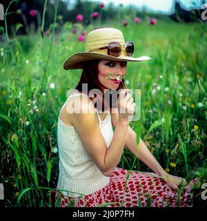 Vintage Italy 1970s, hübsches Frauenporträt mit Sonnenbrille auf Strohhut auf Wiese, riecht nach einer Blume, Europa, Stockfoto