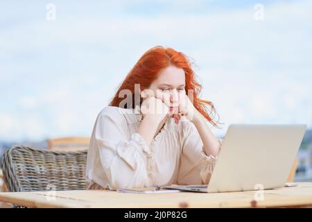 Gelangweilte Frau, die im Café auf der Straße sitzt und aus der Ferne am Laptop arbeitet. Rothaarige Hündin Stockfoto