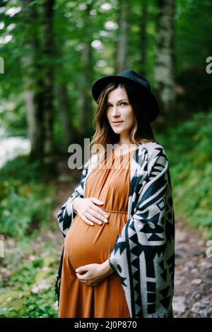 Schwanger Frau in Hut umarmt Bauch mit Händen im grünen Park Stockfoto