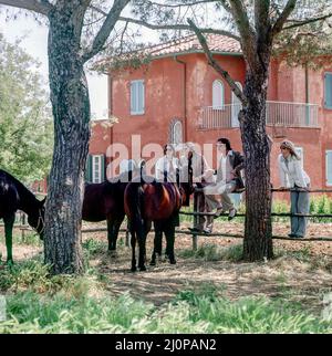 Jahrgang Italien 1970s, 2 Paare von Freunden besuchen Pferdezucht, sitzen am Zaun, römische Landfarm, rosa Haus, Latium, Europa, Stockfoto