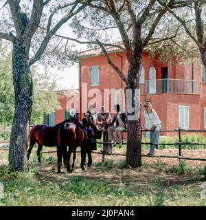 Jahrgang Italien 1970s, 2 Paare von Freunden besuchen Pferdezucht, sitzen am Zaun, römische Landfarm, rosa Haus, Latium, Europa, Stockfoto