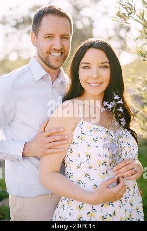 Der Mann umarmt die Schwangere mit Wildblumen an den Schultern Stockfoto