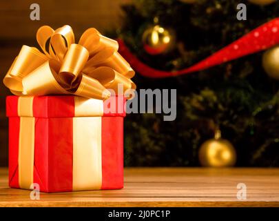 Neujahrsgeschenk in rotem Geschenkpapier mit einer großen goldenen Schleife und Goldriegel. Tannenbaum Hintergrund mit goldenen Kugeln. Speicherplatz Kopieren. C Stockfoto