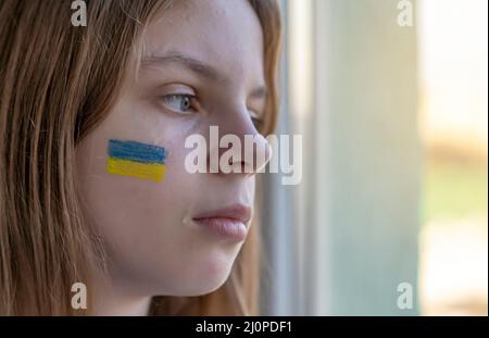 Krieg. Russlands Invasion der Ukraine. Porträt eines Kindes mit Tränen in den Augen, das auf dem Tisch sitzt und aus dem Fenster schaut. Das Blau und Gelb Stockfoto