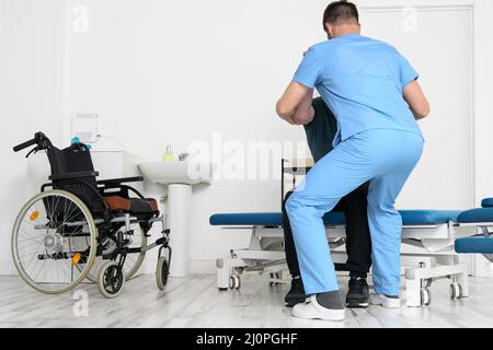 Männlicher Physiotherapeut, der einem Patienten mit einer Behinderung, der einen Rollstuhl benutzt, hilft, im Reha-Krankenhaus aufzustehen. Stockfoto