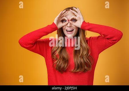 Waist-up-Aufnahme von kindisch und verspielt lustige Ingwer Mädchen in rot gestrickt Kleid macht Gesichter zeigen Kreise über den Augen wie eine Brille Stockfoto