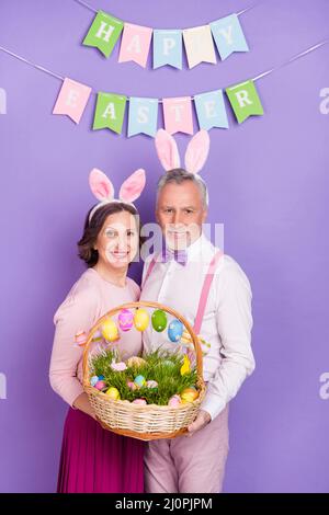 Vertikale Foto von zwei älteren fröhlichen Menschen halten Eier Korb Hasenohren isoliert auf violettem Hintergrund Stockfoto