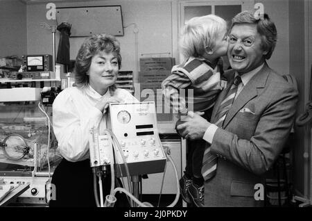 Der Fernsehpersönlichkeit Michael Aspel, der Vizepräsident von Bliss ist, kehrte ins West London Hospital zurück, um einige Geräte an die Special Care Baby Unit zu übergeben. Mit ihm waren seine Frau Lizzie Power und der 3-jährige Sohn Patrick, der vorzeitig geboren wurde und viele Wochen in der Babyeinheit verbrachte. Die von der Wohltätigkeitsorganisation gespendete Ausrüstung ist ein Beatmungsgerät – ein unverzichtbarer Bestandteil, um Neugeborenen zu helfen, die alleine nicht atmen können. Es wurde dem beratenden Kinderarzt Dr. H Barry übergeben. 3.. Oktober 1984. Stockfoto