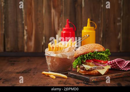 Hausgemachter Burger mit Rind-, Käse- und Zwiebelmarmelade auf einem Holzbrett, Pommes in einem Metallkorb und Saucen. Fast-Food-Konzept, Stockfoto