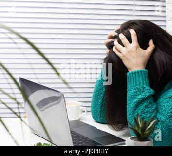 Eine Frau in einem Pullover sitzt am Tisch mit dem Kopf in den Händen, Stress und Kopfschmerzen durch Überlastung, Müdigkeit. Ein Notebook Stockfoto