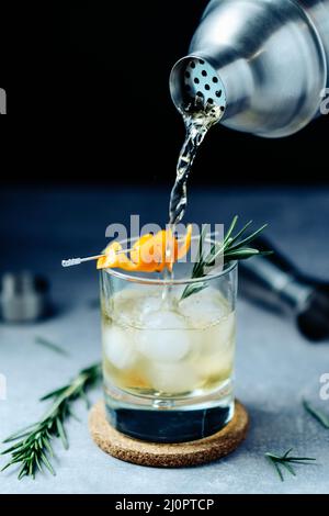 Alkoholcocktail aus dem Shaker in Glas mit Orangenschale Rosmarin und Eis auf Holztisch Stockfoto