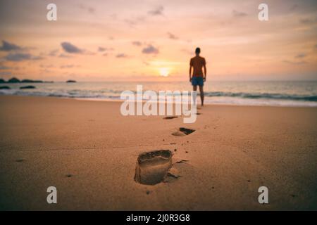 Fußabdrücke im Sand gegen die Silhouette der Person. Einsamer Mann, der bei goldenem Sonnenuntergang am Strand entlang zum Meer geht. Stockfoto